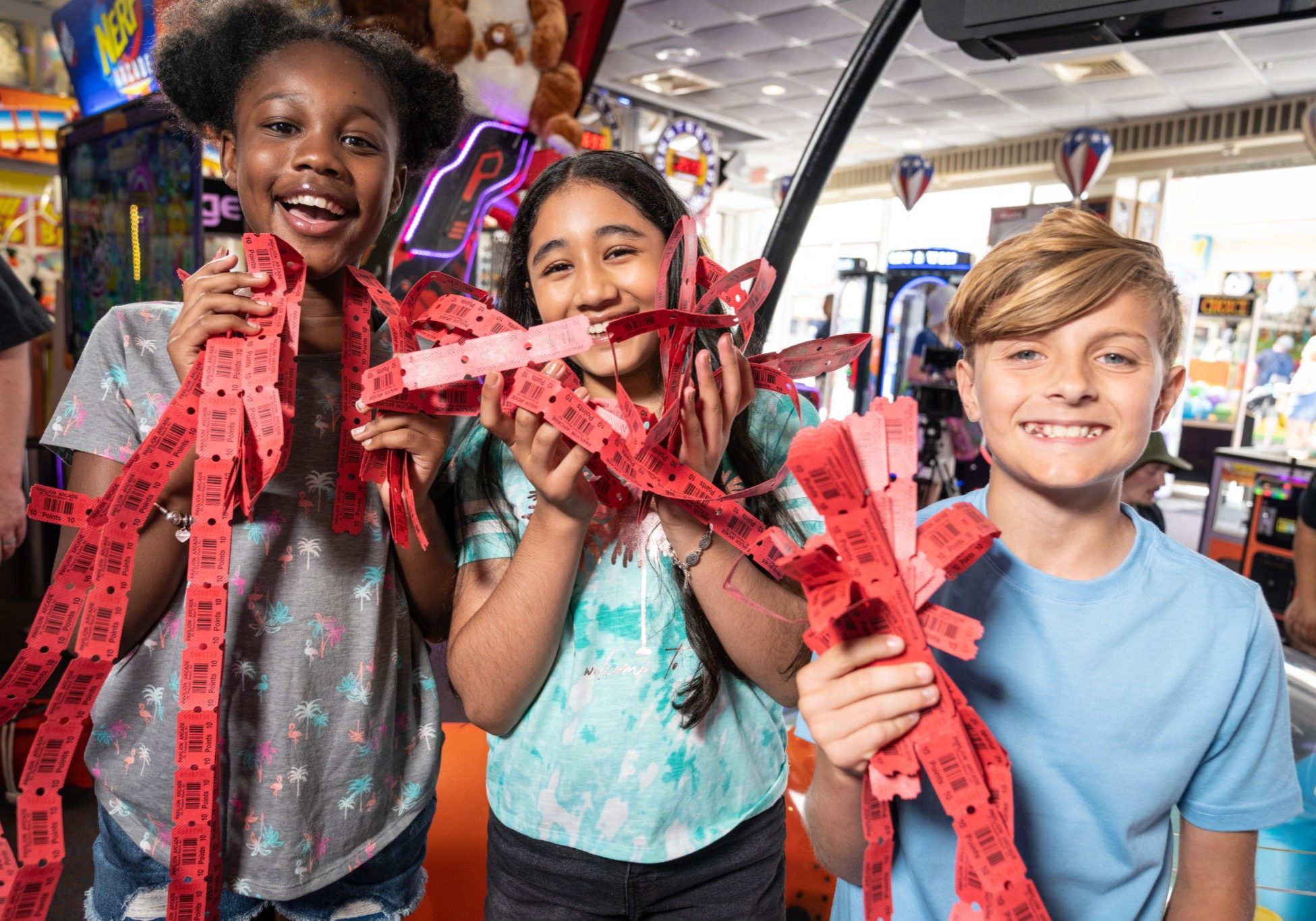 Three children smiling and holding up a colorful ribbon at a lively carnival event.